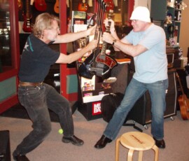 Two men mock fighting with guitars indoors.